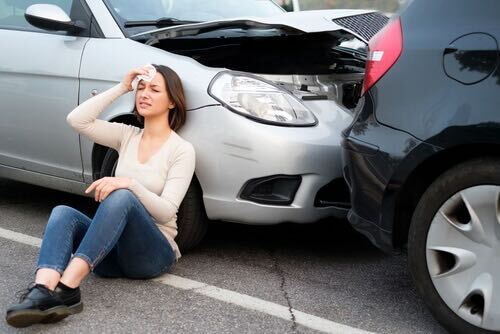 Woman sitting on the ground beside a damaged car, holding her head, illustrating the need for auto injury transportation services in Manchester, MI.