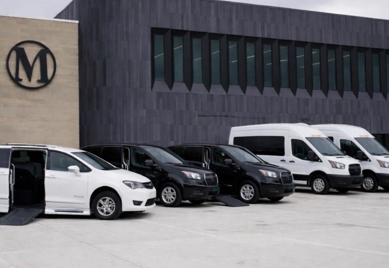 White and black wheelchair accessible vehicles parked outside a modern building with the MBM Transportation logo, showcasing safe patient transportation options in Ferndale.