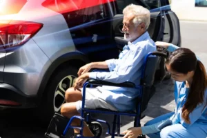 Elderly man in a wheelchair assisted by a caregiver near a parked car.